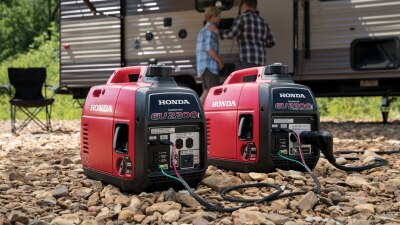 Deux génératrices Honda montrées ensemble sur une surface rocheuse devant deux personnes jasant à l’extérieur d’une caravane.