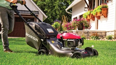 Une personne en jeans pousse une tondeuse devant une propriété.  