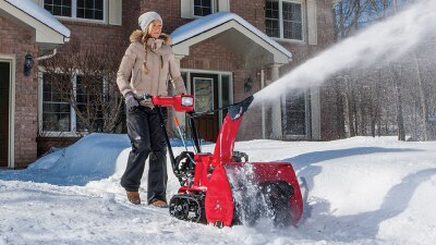 Plan étendu d’une femme déblayant l’entrée avec une souffleuse à neige par une journée ensoleillée. Derrière elle se trouve une belle maison de prestige en briques rouges.