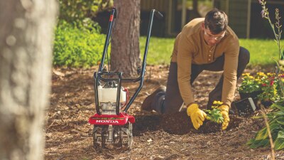 Un jardinier, à genoux, plante des fleurs derrière un motoculteur Honda. 