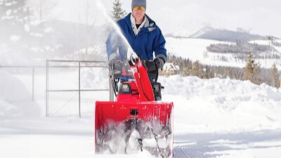 Front-facing image of man in blue jacket pushing red honda snowblower. Snow landscape in background. Trees and mountains in the distance.