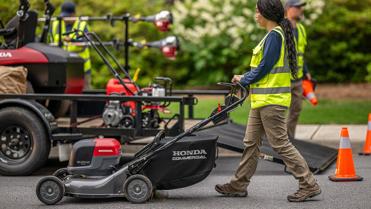 Worker pushing the battery lawn mower on the street next to a trailer. 
