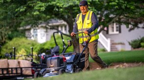 View from three-quarter front left showing a worker pushing a battery lawn mower on the lawn. 