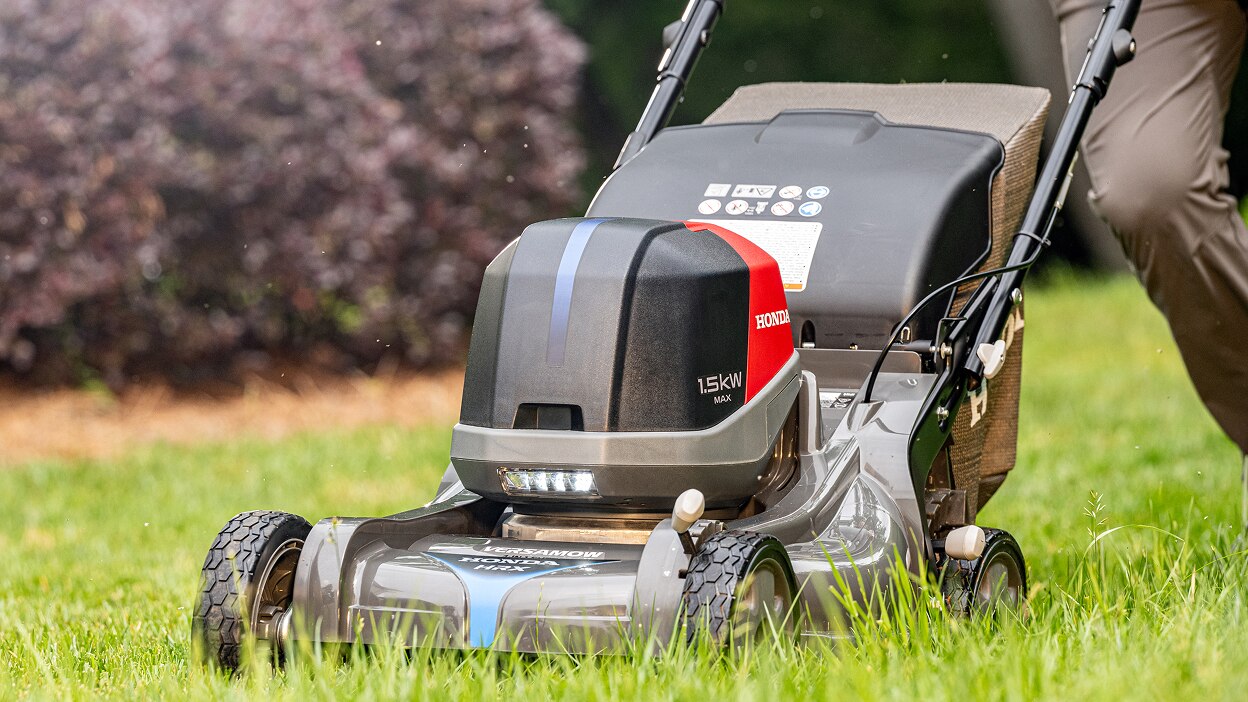 View from three-quarter front left showing a person pushing a battery lawn mower to mow the lawn. 