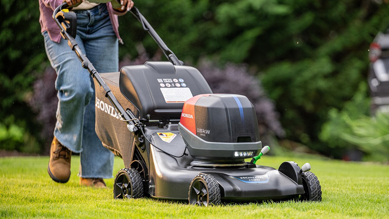View from three-quarter front showing a person pushing a battery lawn mower to mow the lawn. 