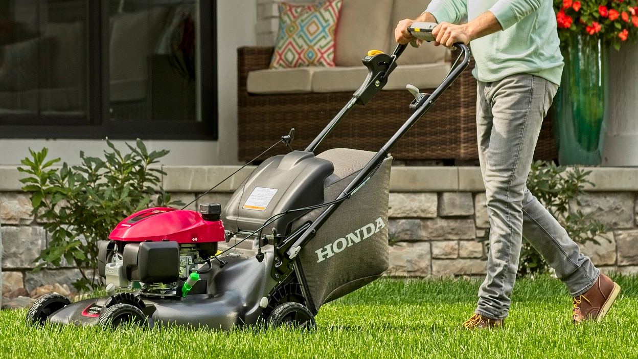 Person mowing lawn going left. Small brick wall in background.