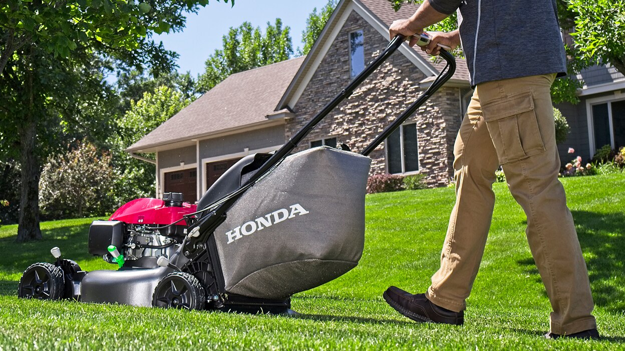 Une personne tond la pelouse en allant vers la gauche. Une maison en arrière-plan.
