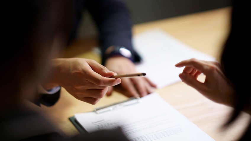 Close-up of a person passing a pen to another person who has documents on the table in front of them. The edges of the photo are blurry, with the emphasis only on swapping the pen between the hands.