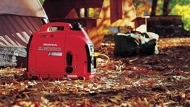 Gros plan au niveau du sol d'une génératrice Honda EU 1000i rouge posée sur le sol feuillu d'un camping à côté d'une tente grise, avec un sac de sport vert et une table de pique-nique en bois en arrière-plan.